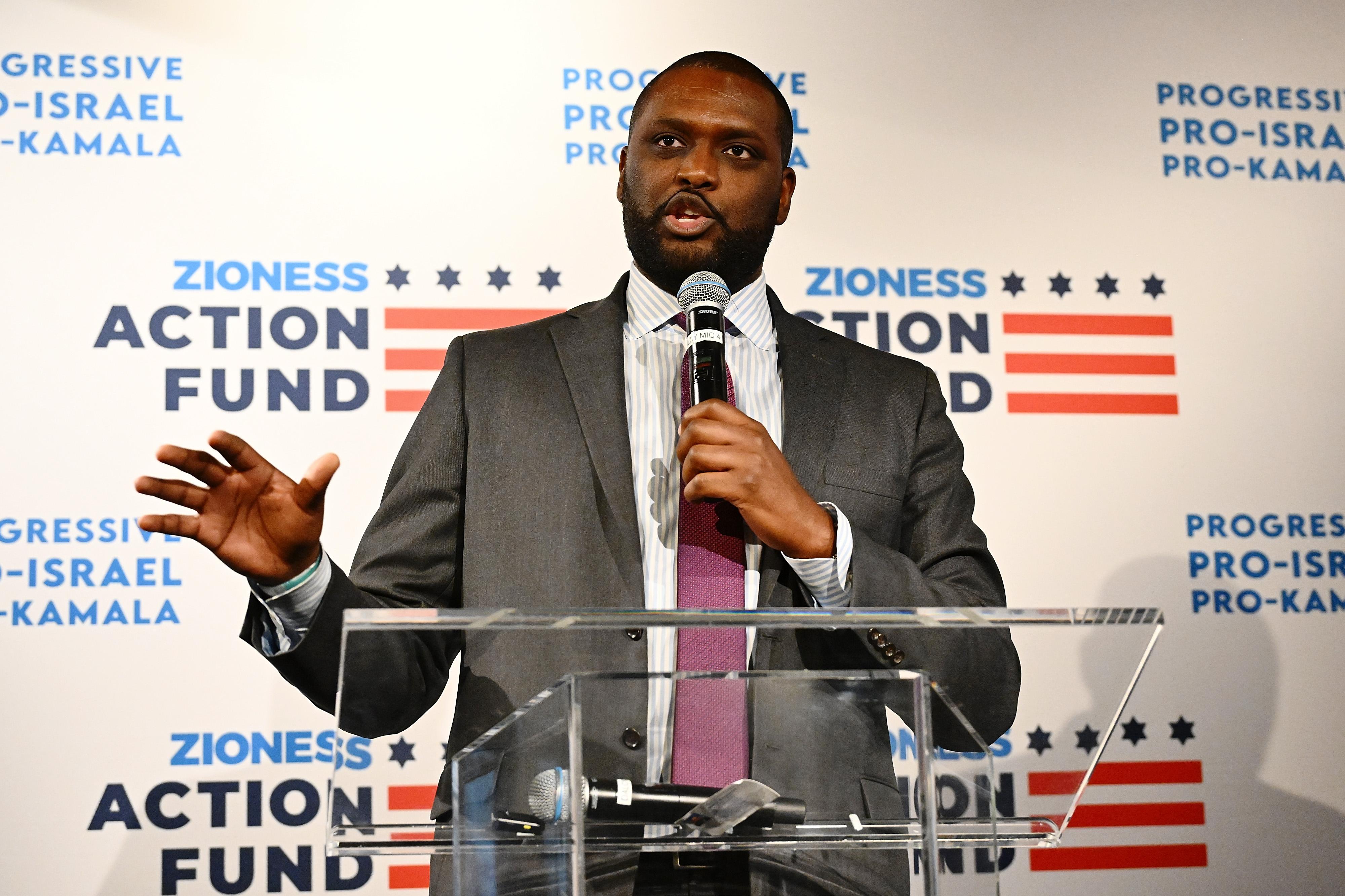 A bearded Black man holds a mic and stands at a lectern, speaking. Behind him is a background that reads "Zioness Action Fund."