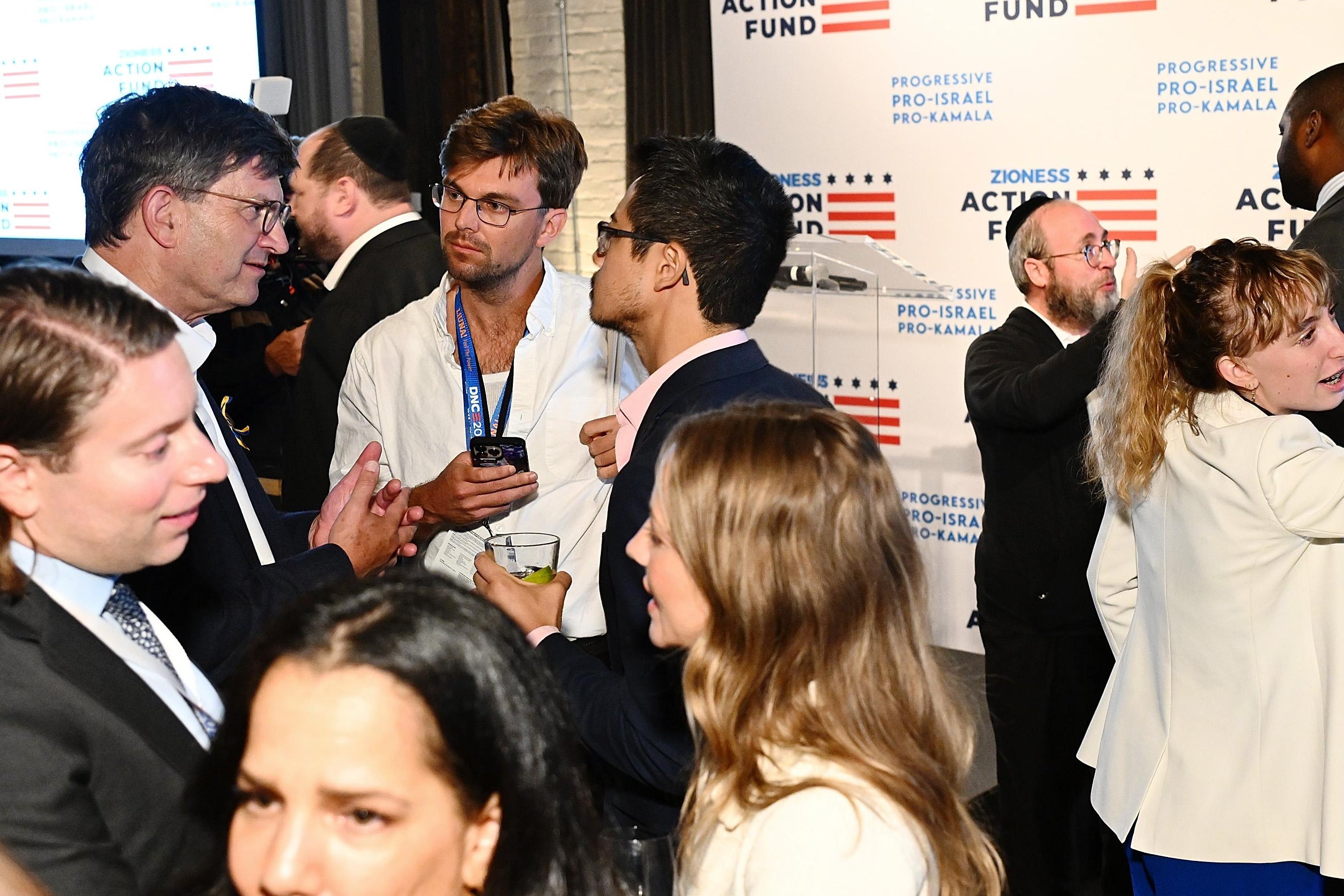 People in suits speaking to each other in front of a backdrop that says "Zioness Action Fund: Progressive, Pro-Israel, Pro-Kamala."