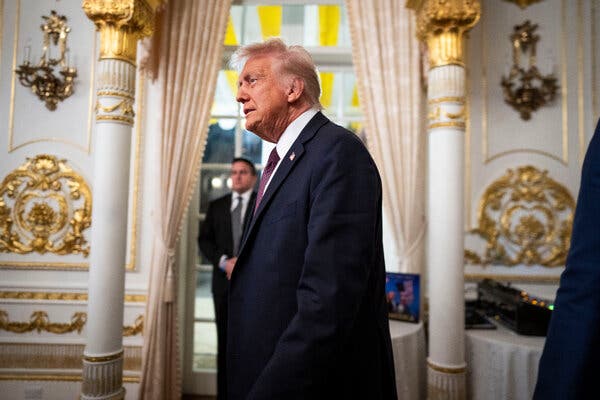 Donald J. Trump, in a dark suit and burgundy tie, is seen from the side in an ornate gold and white room.