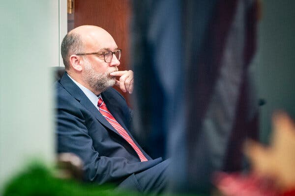 Russell Vought, wearing a suit and a red striped tie, sits in a chair with an index finger raised to his lips. 