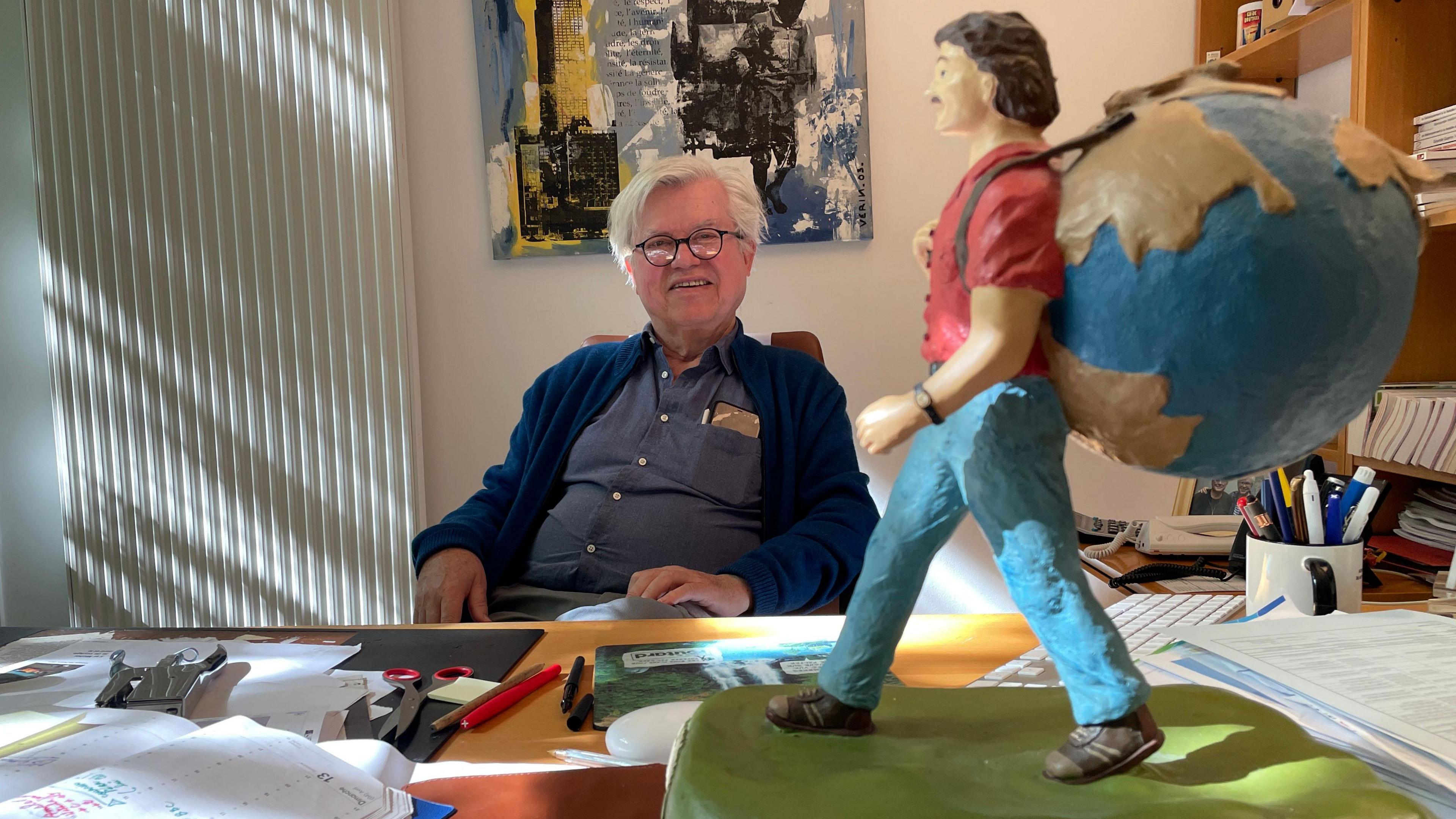 Philippe Gloaguen, the founder of French travel guides, Le Guide du Routard, is seen sitting at a desk. In the foreground is a model of a backpacker carrying the globe.