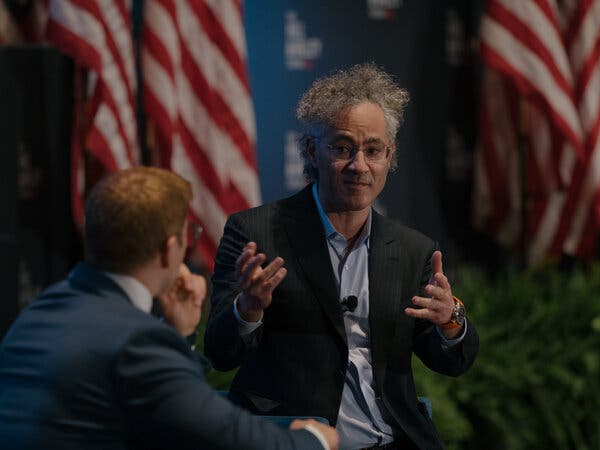 Alex Karp in a dark jacket and collared shirt, gesturing with his hands.