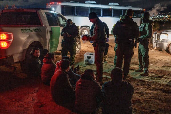 A nighttime image of four people standing in front of a group of people who are sitting on the ground; next to them is a vehicle displaying the words Border Patrol. 