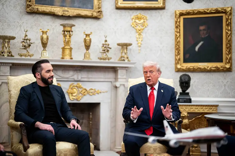 Two men wearing navy suits, one with dark hair and no tie, the other with light hair and a large red tie, sit in armchairs in a room decorated with gilded vases and ornamentation.