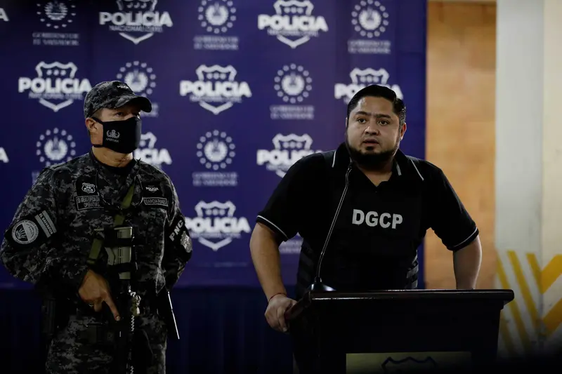 A man in a polo shirt at a speaking podium is flanked by a masked man in camouflage holding an automatic weapon, in front of a step-and-repeat backdrop depicting logos for the local police agency.