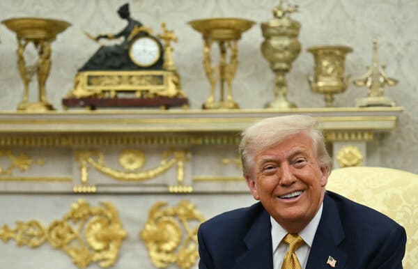 President Trump, in a gold tie, smiles in front of an Oval Office mantel covered in gold decoration and gold decorative objects.