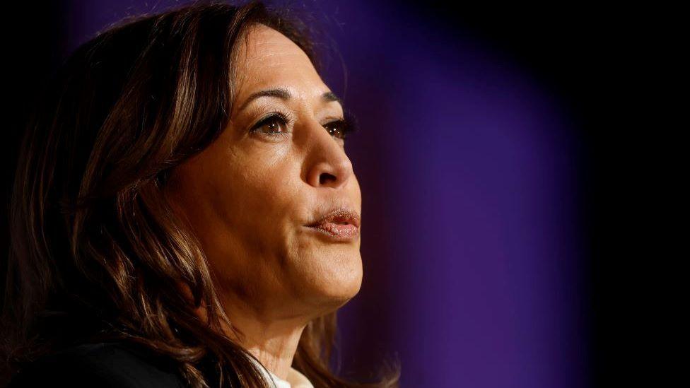 Kamala Harris looks to the right of the camera, wearing a suit and her back lit in mauve light