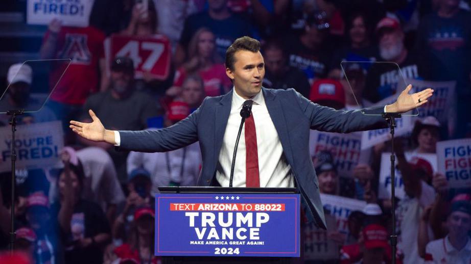 An image of Charlie Kirk speaking at a Trump rally in Glendale, Arizona in August 2024. Behind him is a crowd donned in red, white and blue clothing, holding signs that say 'YOU'RE FIRED' and 'FIGHT! FIGHT! FIGHT!'. He is wearing a blue suit with a white button-up top and a red tie, speaking at a podium while gesturing widely with his hands. On the podium is a sign that reads 'TRUMP-VANCE MAKE AMERICA GREAT AGAIN 2024'. 