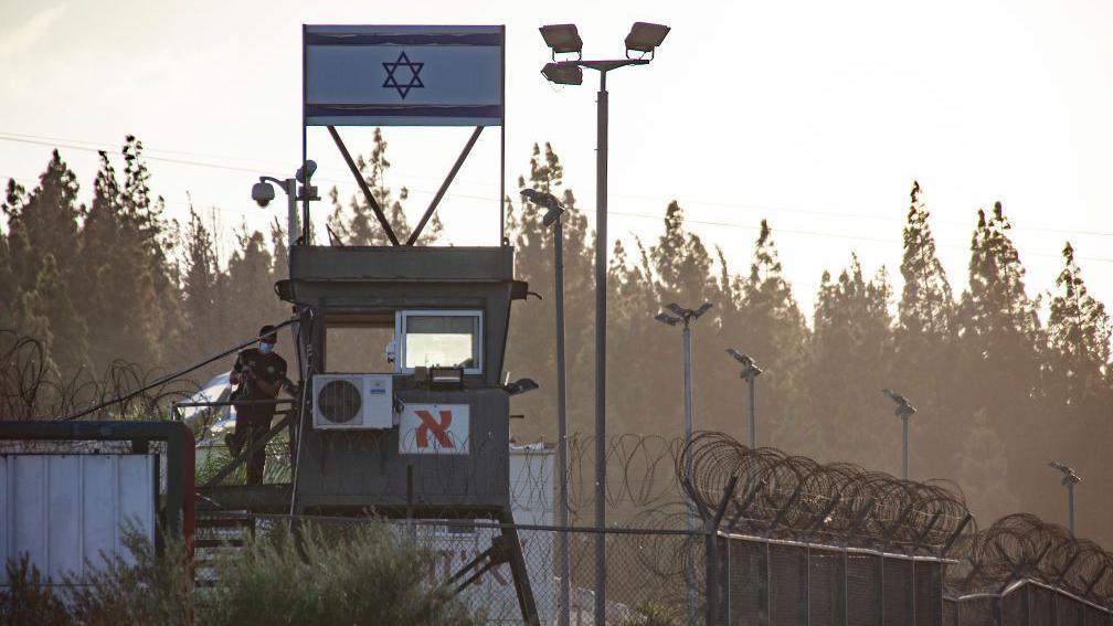 A file photograph of Megiddo prison in Israel shows a watchtower with an Israeli flag above it. Coiled barbed wire can be seen on top of high fences, with a line of trees in the background. 