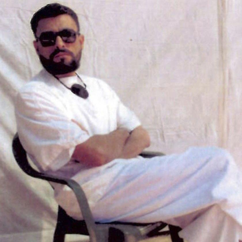 Abu Zubaydah wearing a white t-shirt and white trousers, carrying an eye-patch around his neck. He is also wearing black sunglasses and has a defined beard.
