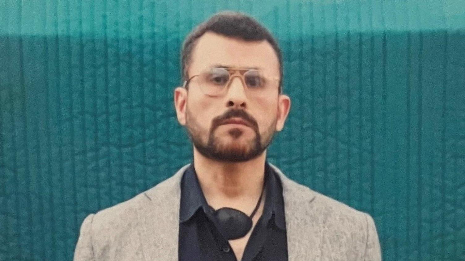 A head-to-chest shot of Abu Zubaydah, a man wearing glasses and with a defined beard, carrying an eye-patch around his neck. He is wearing a black shirt and grey blazer.