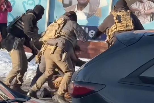 A group of officers wearing masks and camouflage vests surround a man who is crouched on the ground.
