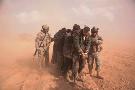 Two US soldiers escort four or five blindfolded and handcuffed men across sandy ground