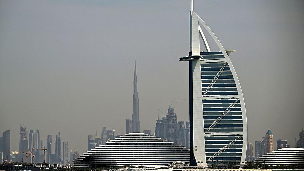 Buildings on the Dubai skyline, with tall towers seen in the background and the sail-shaped hotel the Burj Al Arab in the foreground, against a grey sky 