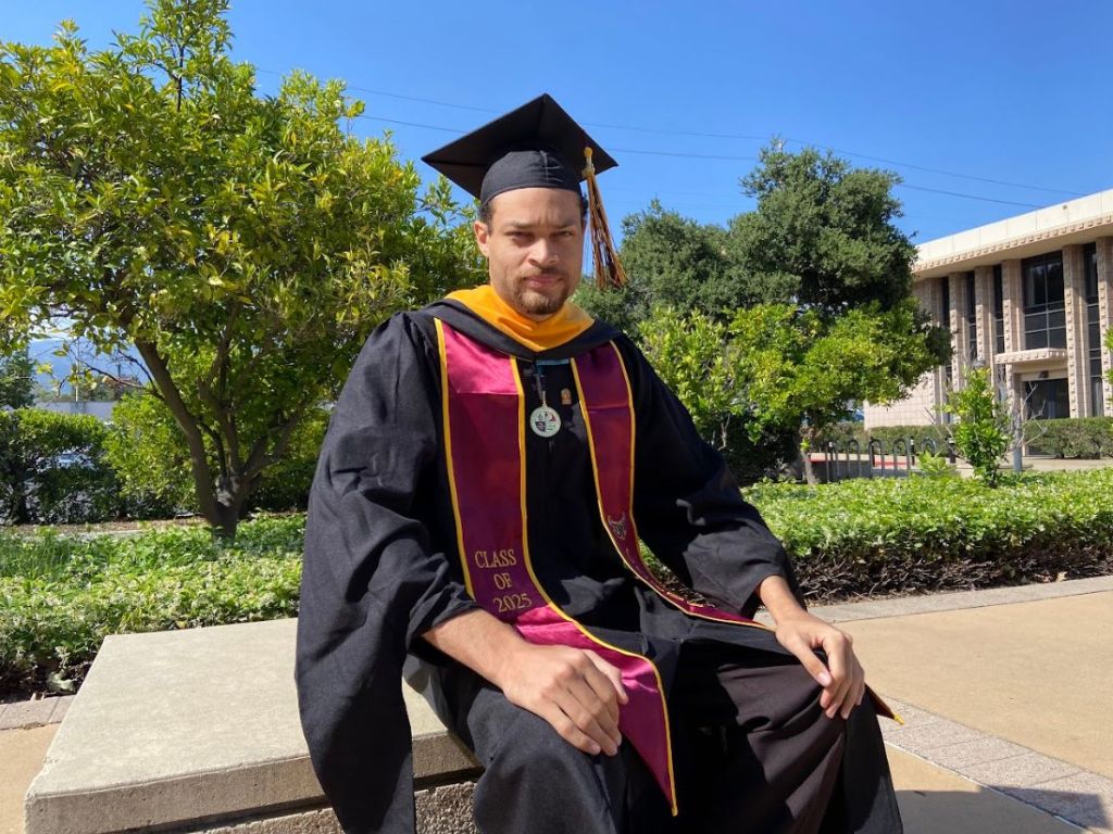 A man in a graduation cap and gown with a "Class of 2025" stole sitting outdoors.