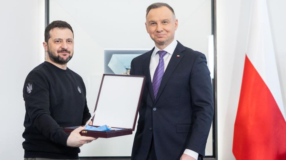 President Volodymyr Zelensky, bearded and wearing a black jumper, holds a box containing a medal next to then-President Andrzej Duda, who is wearing a suit and stands next to a red and white flag.