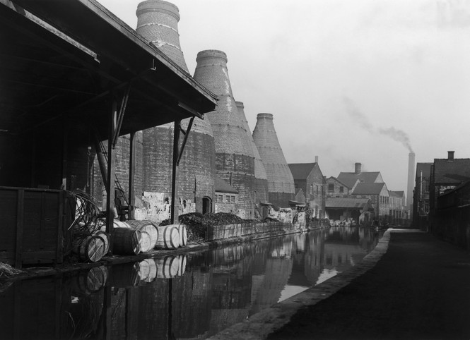 black-and-white photo of large bottle-shaped buildings along canal, with homes and smokestack in background