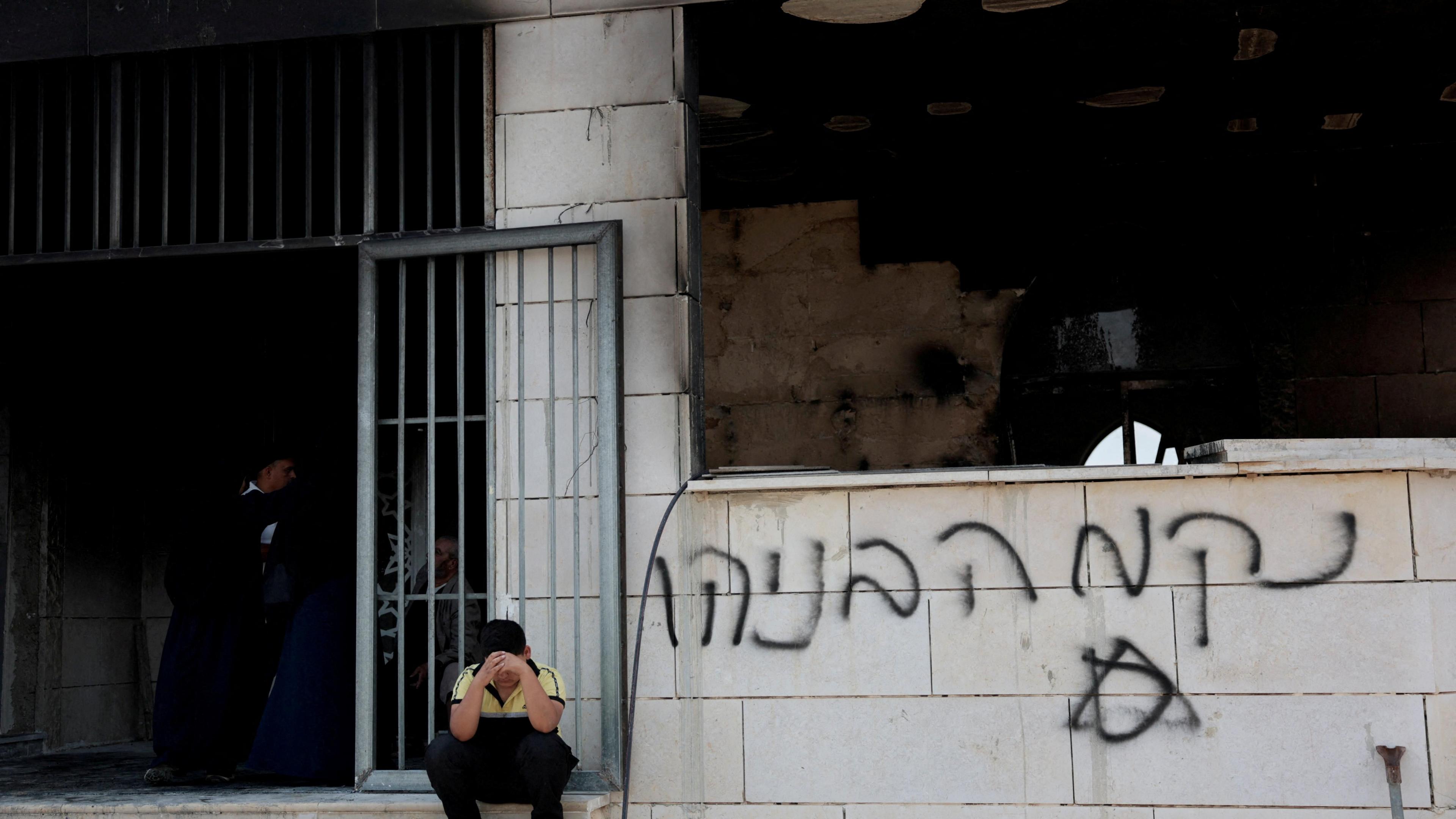 A boy sits with his face in his hands outside a damaged mosque which has graffiti on its walls