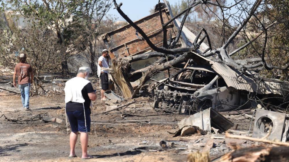 Jewish settlers, wearing religious garments, stand around the remains of a burnt building and car, in Havat Gilad (19/07/26)