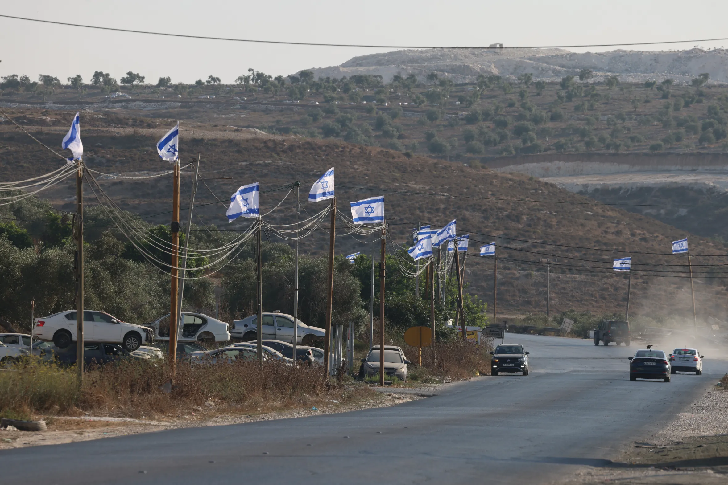 Israeli flags flown from poles along a road leading to Jenin City, with cars parked on the side and a hill in the background.