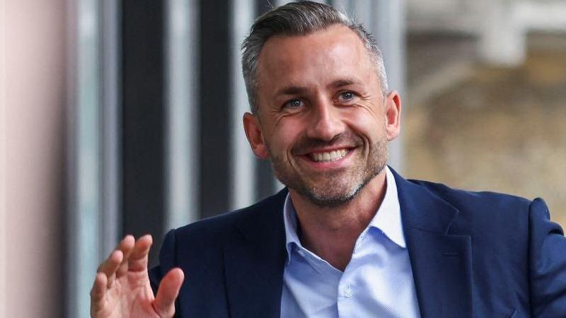 Ulrich Siegmund, who has greying hair and a short beard and wears a navy suit jacket with a light blue shirt, smiles and waves as he arrives at a press conference on Monday