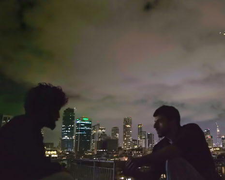 A man's silhouette in profile sits on a rooftop overlooking a lit city skyline at night