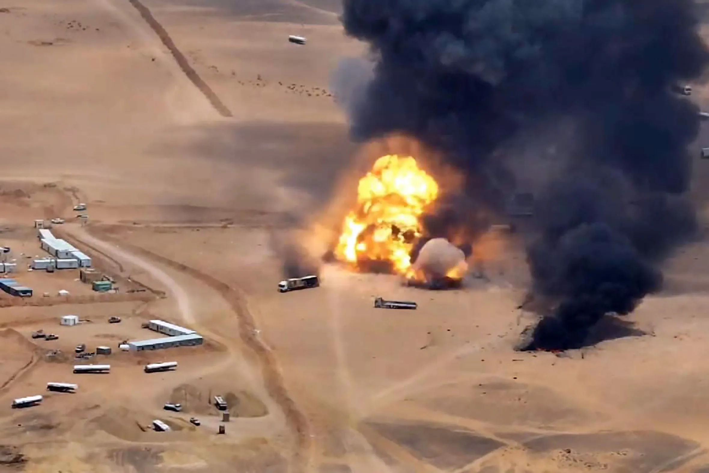 An aerial view of a massive explosion and thick black smoke billowing over a desert landscape, near a compound of buildings and several trucks.